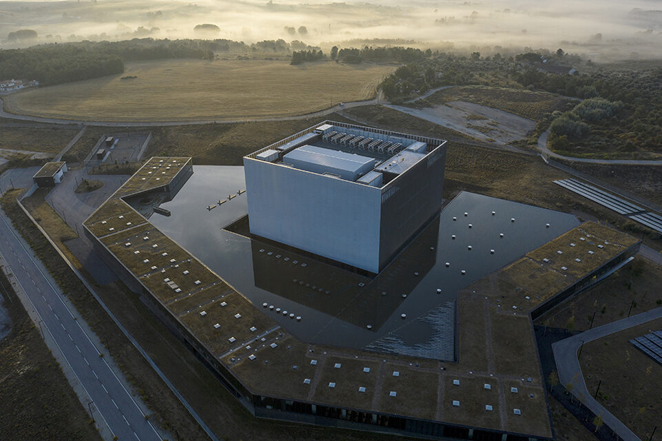 Data centers contribute about the same amount of greenhouse gas emissions as the aviation sector, according to the United Nations. Climate change activists have been pushing providers of digital services to reduce electricity use, especially as the sector booms in an era of Big Data.
Portugal’s Covilhã Data Center, one of the largest in Europe, doesn’t just look cool. It was designed to stay cool—even if deadly heat waves like the one that hit Europe this summer become more frequent. The brutalist facility is spread over 75,500 square meters, an area equivalent to about 10 soccer fields; it’s big enough to house as many as 56,000 servers. 
The owner, Altice Portugal, the biggest telecommunications services provider in the country and a subsidiary of Altice Europe NV, says the five-year-old center was built with efficiency in mind.
Its location, in Covilhã, a ski and hiking destination 170 miles northeast of Lisbon, was chosen partly because of the mild mountain climate. Outside air cools the building’s servers 99 percent of the time, according to the company. The center also collects and stores rainwater, creating a sustainable water source—and surrounding its campus with a striking moatlike feature.